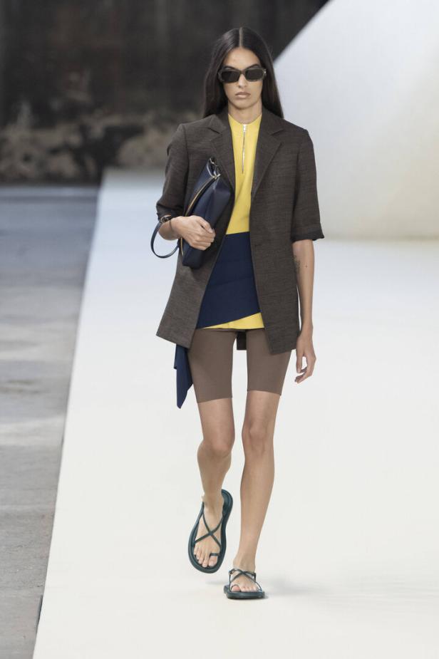Eine Frau mit Sonnenbrille läuft in einem braunen Blazer und Radlerhosen über einen Laufsteg.