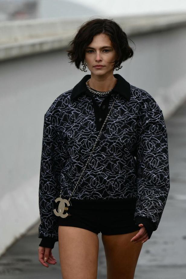 Eine Frau mit kurzem, dunklem Haar trägt ein Outfit von Chanel mit einer goldenen Handtasche.