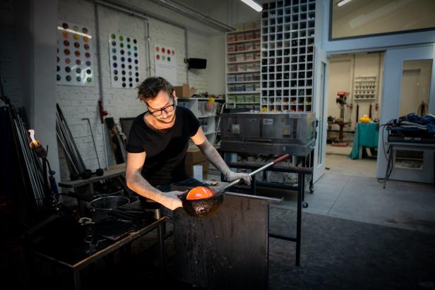 Ein Glasbläser formt in seiner Werkstatt eine glühende, orangefarbene Kugel.