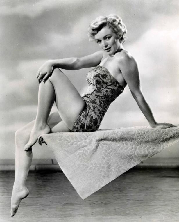Die amerikanische Schauspielerin Marilyn Monroe posiert in einem Badeanzug auf einem Sprungtuch sitzend.