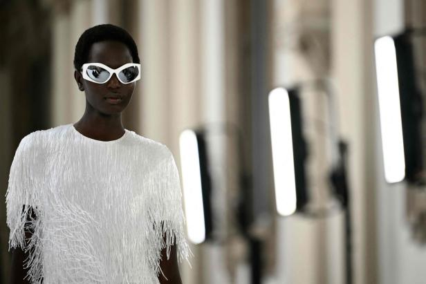 Eine Frau mit weißem Kleid und futuristischer Sonnenbrille auf einer Modenschau.