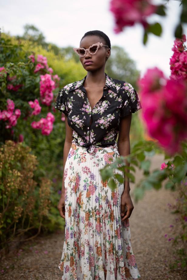 Eine Frau mit floral gemustertem Hemd und Rock steht in einem Garten.