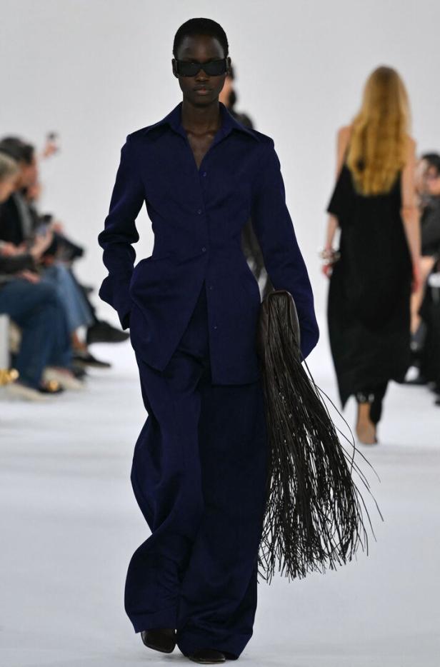 Eine dunkelhäutige Frau in einem marineblauen Hosenanzug mit einer Fransenhandtasche auf dem Laufsteg.