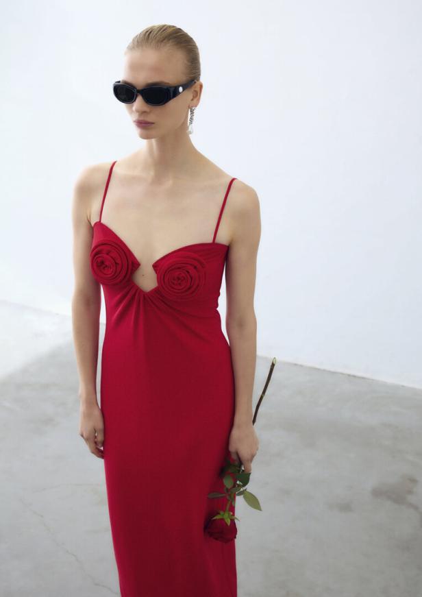 Eine Frau mit Sonnenbrille trägt ein rotes Kleid mit Rosenapplikationen und hält eine Rose.