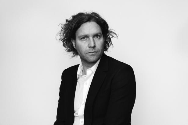 Ein Mann mit lockigem Haar trägt ein schwarzes Sakko und ein weißes Hemd.