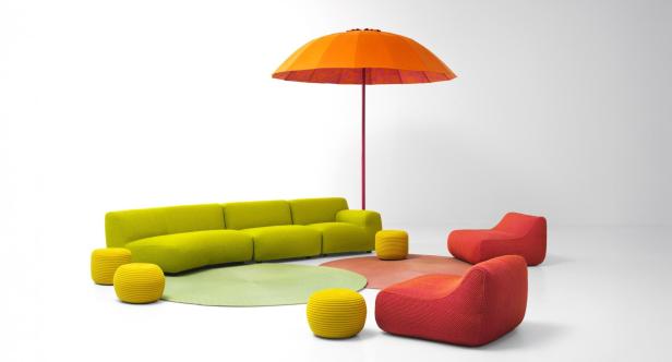 Ein Arrangement aus grünen Sofas, roten Sesseln, gelben Hockern und einem orangefarbenen Sonnenschirm.