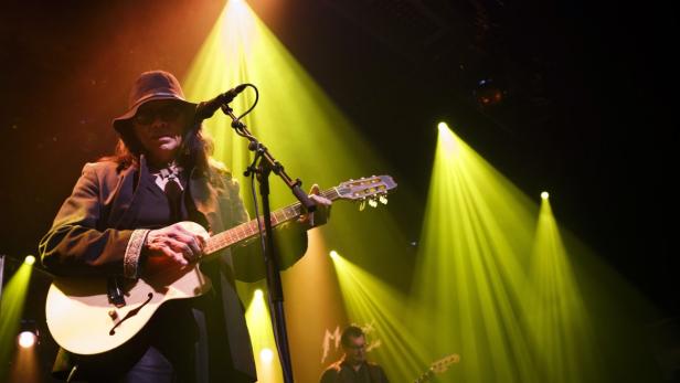 Ein Musiker mit Hut spielt Gitarre auf einer Bühne mit gelbem Scheinwerferlicht.