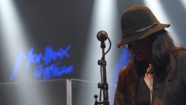 Ein Musiker mit Hut steht auf der Bühne vor einem Mikrofon.