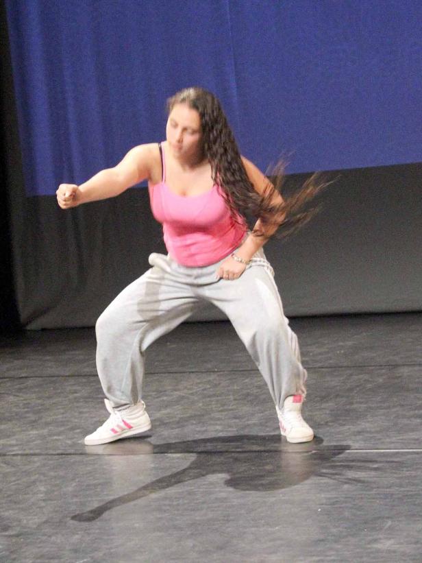 Eine junge Frau tanzt in einem rosa Top und grauer Jogginghose auf einer Bühne.