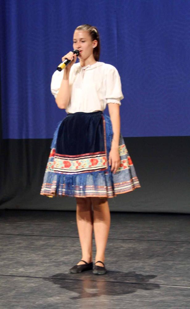 Ein junges Mädchen singt in traditioneller Tracht auf einer Bühne.