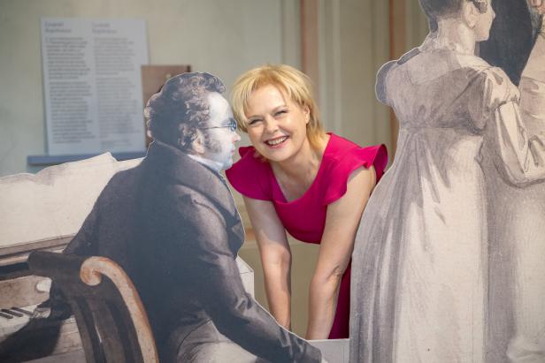 Eine Frau in einem pinkfarbenen Kleid posiert mit Pappaufstellern von Franz Schubert und einer anderen Person.