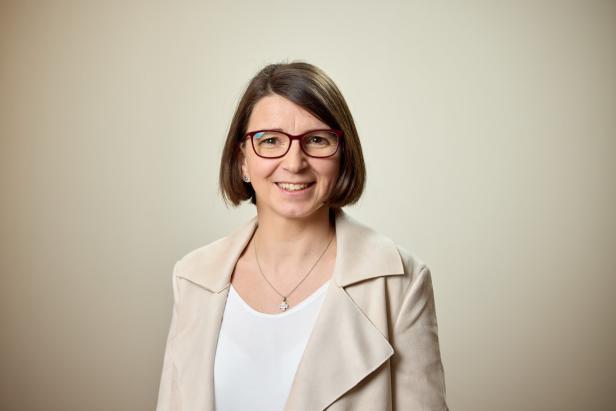 Eine lächelnde Frau mit Brille und beigem Blazer vor einem hellen Hintergrund.