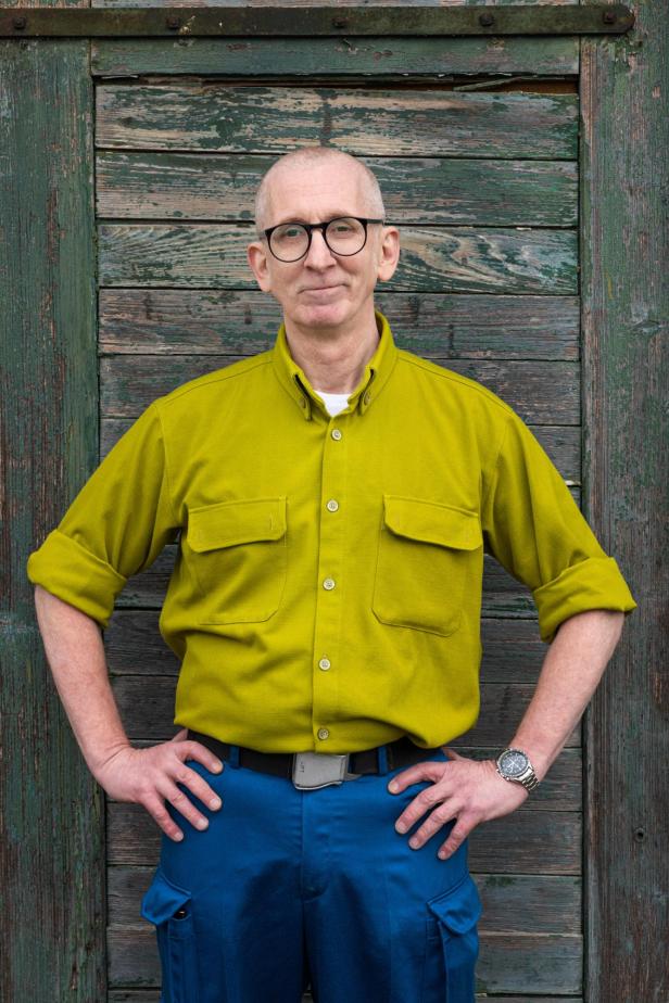 Ein Mann in einem senfgelben Hemd und einer blauen Hose vor einer Scheune.