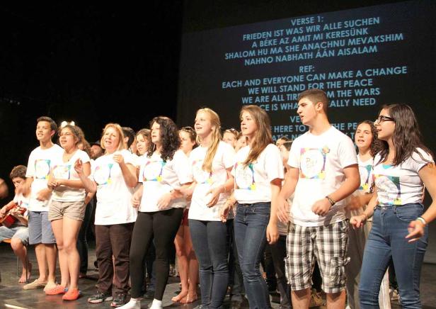 Eine Gruppe junger Leute singt auf einer Bühne vor einem Liedtext, der auf eine Leinwand projiziert wird.