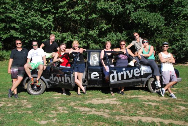 Eine Gruppe von Leuten posiert an und in einem alten, schwarzen Auto mit der Aufschrift „doubt“ und „driver“.