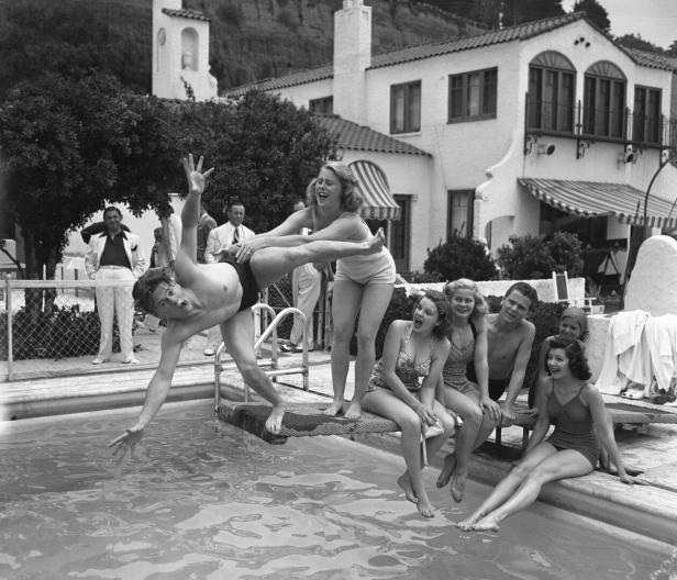 Junge Menschen sitzen auf einem Sprungbrett. Ein Mädchen wirft einen jungen Mann ins Wasser. Alle lachen.