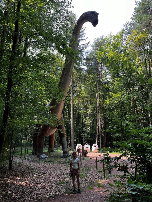 Ein Mädchen steht vor einem riesigen Dinosaurier-Modell im Wald.