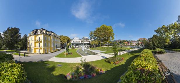 Ein Panoramablick auf einen Park mit Springbrunnen und verschiedenen Gebäuden an einem sonnigen Tag.