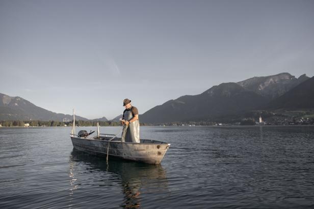 Ein Fischer steht in einem Boot auf einem See vor einer Bergkulisse.