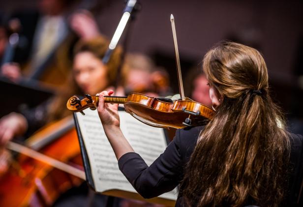 Eine Frau spielt Violine. Hinter ihr ist der Anfang eines Orchesters zu sehen.
