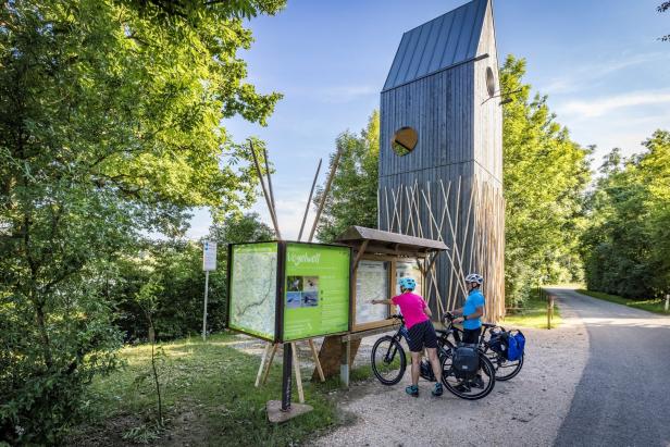 Zwei Radfahrer betrachten eine Informationstafel neben einem hölzernen Aussichtsturm.