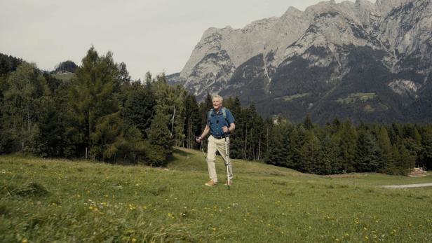 Ein älterer Mann wandert mit Stöcken auf einer grünen Wiese vor einer Bergkulisse.