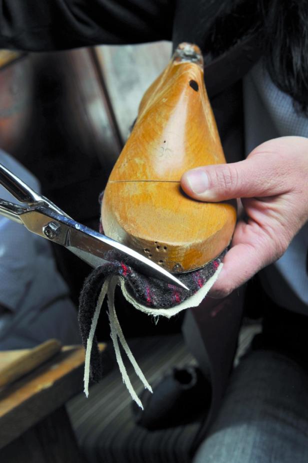 Ein Schuhmacher schneidet Stoff mit einer Schere um einen hölzernen Schuhleisten.