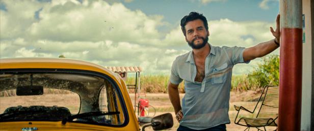 Ein Mann lehnt an einer Säule neben einem gelben Auto vor einer ländlichen Szene.