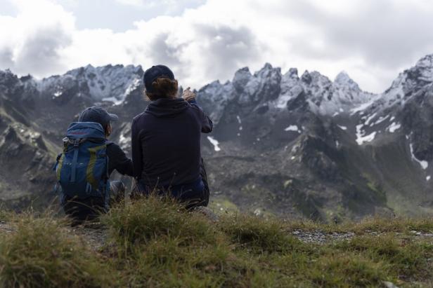 Ein Kind mit Rucksack und eine erwachsene Person sitzen und schauen auf schneebedeckte Berge.