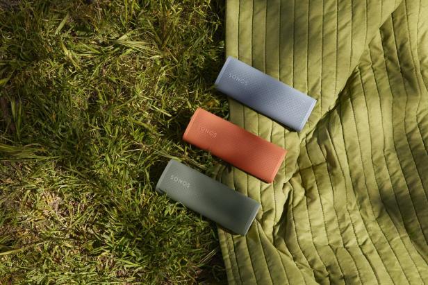 Drei Sonos Roam Lautsprecher in Blau, Orange und Grün liegen auf einer grünen Decke im Gras.