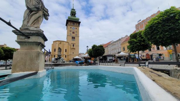 Ein Brunnen auf einem Marktplatz, dahinter ein gelber Turm mit einer grünen Spitze.