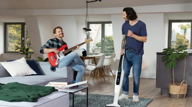 Zwei Männer in einem gemütlichen Wohnzimmer, einer spiel Gitarre der andere staubsaugt..