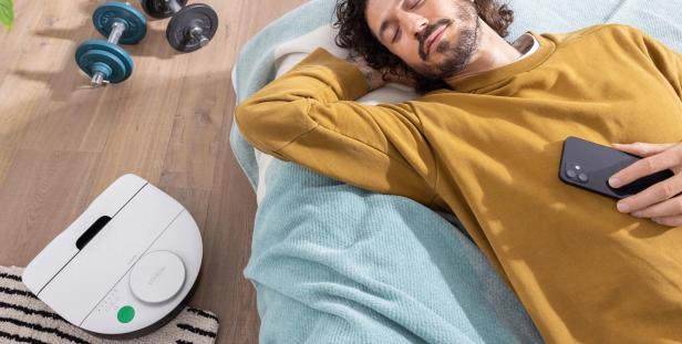 Ein Mann schläft im Bett mit seinem Handy in der Hand und ein Saugroboter reinigt den Boden.