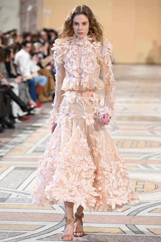 Ein Model präsentiert ein elfenbeinfarbenes Kleid mit floralen Applikationen auf einem Laufsteg.