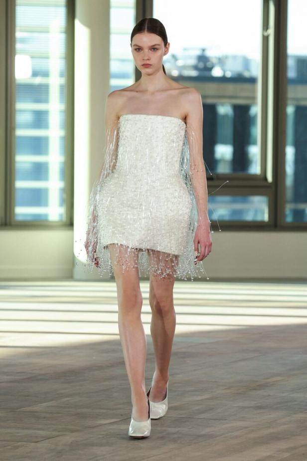 Ein Model präsentiert ein trägerloses, weißes Minikleid mit Fransen auf einem Laufsteg.