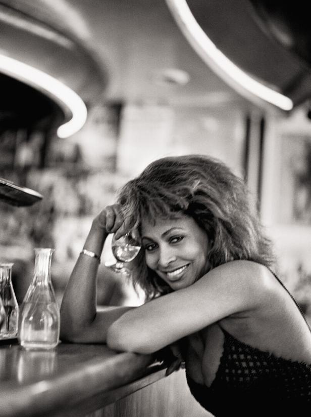 Tina Turner lehnt an einer Bar und hält ein Glas in der Hand.