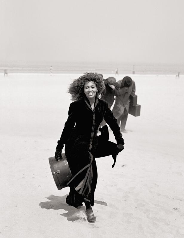 Tina Turner läuft mit einem Koffer am Strand entlang.