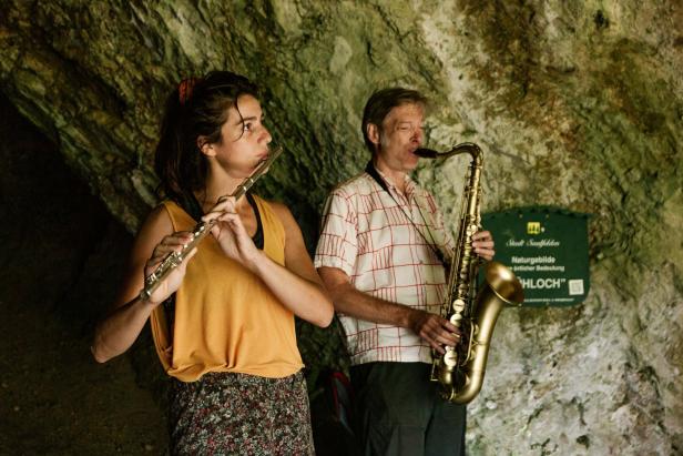 Eine Frau spielt Flöte, während ein Mann Saxophon spielt; beide stehen vor einer Felswand.