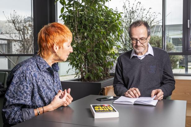 Eine Frau mit rotem Haar spricht mit einem Mann, der in ein Notizbuch schreibt. Auf dem Tisch liegt ein Buch von Eva Rossmann.