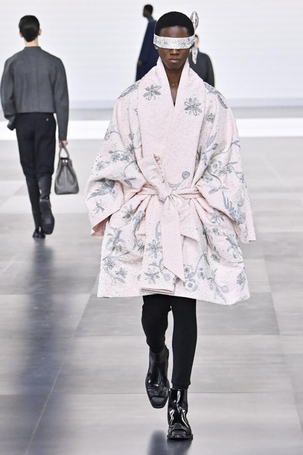 Ein Model mit einer verzierten Augenbinde und einem rosa, blumengemusterten Mantel auf dem Laufsteg.