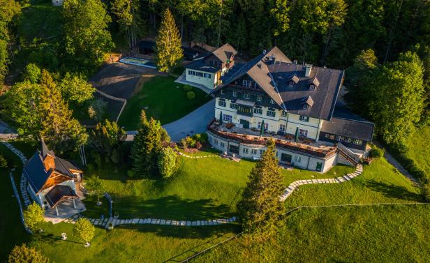 Luftaufnahme eines großen Hauses mit Nebengebäude und Pool auf einem grünen Hügel.