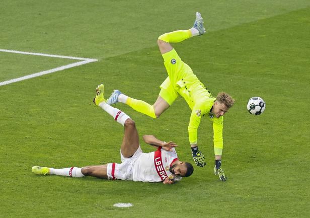 Ein Fußballspieler liegt am Boden, während ein Torwart in leuchtend gelber Kleidung in der Luft ist.