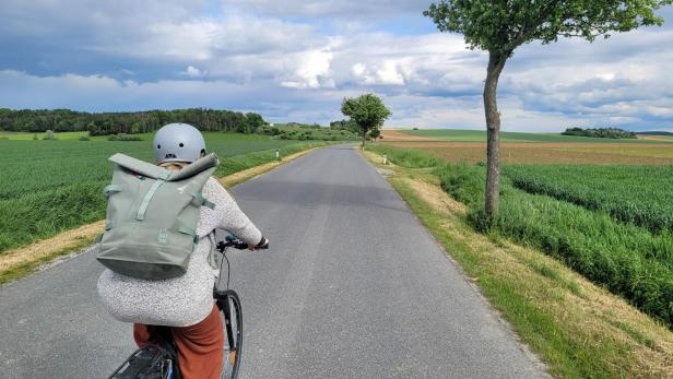 Eine Fahrradfahrerin zwischen grünen Wiesen und Feldern auf einer Landstraße.