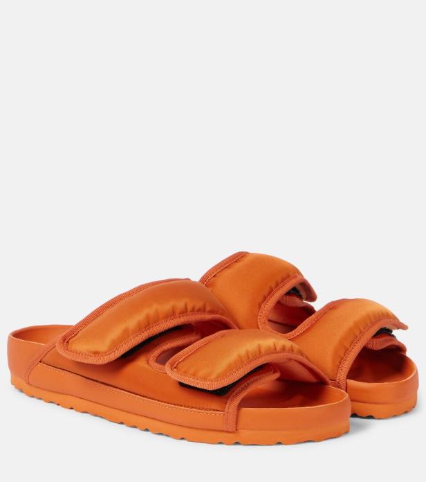 Ein Paar orangefarbene Pantoletten mit verstellbaren Riemen stehen vor einem neutralen Hintergrund.