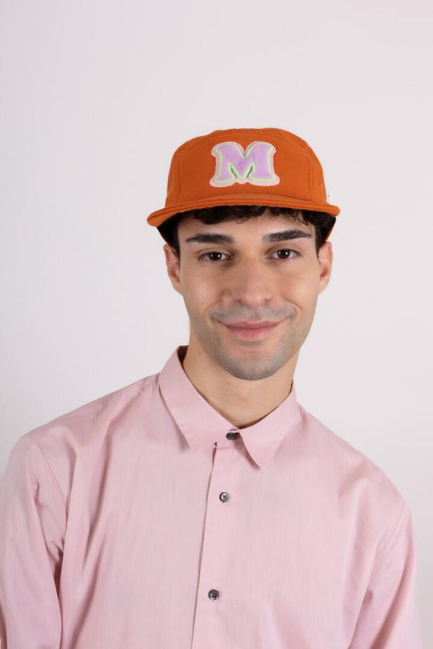 Ein Mann mit einem orangefarbenen Cap mit dem Buchstaben M und einem rosa Hemd.
