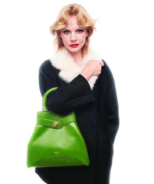 Eine Frau mit blonden Haaren und rotem Lippenstift hält eine grüne Prada-Handtasche.