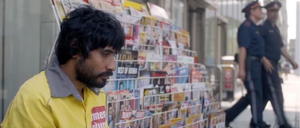 Ein Zeitungsverkäufer steht vor einem Zeitschriftenstand, im Hintergrund Polizisten.