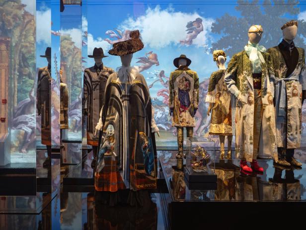 Mehrere Mannequins tragen extravagante, mit religiösen Motiven bedruckte Mäntel vor einem gemalten Himmel.