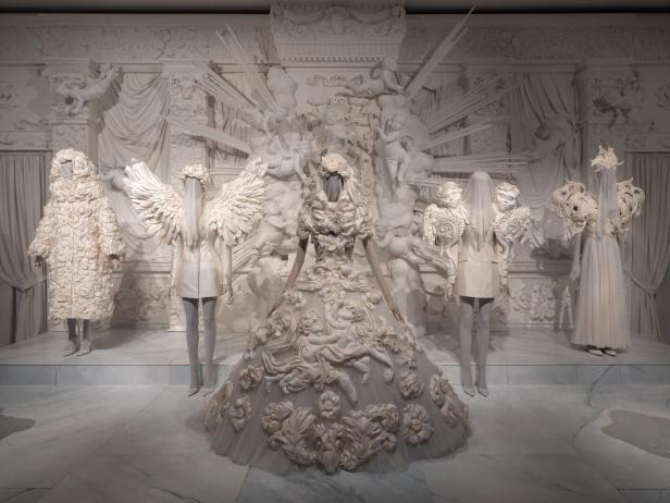 Mehrere weiße Kleider und Kostüme sind in einer Ausstellung mit Engelsstatuen zu sehen.