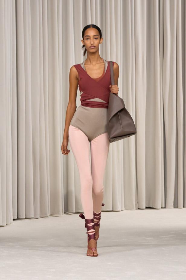 Eine Frau in einem rot-braunen Oberteil, einem beigen Body und rosa Leggings mit einer großen Tasche.